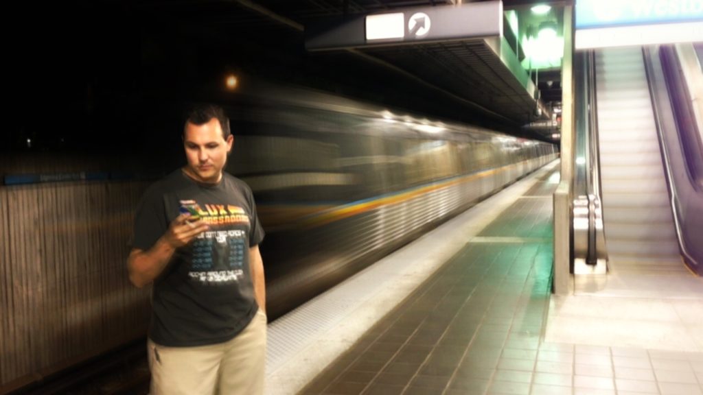 Bill Selak looking at a phone while a train passes, blurred behind